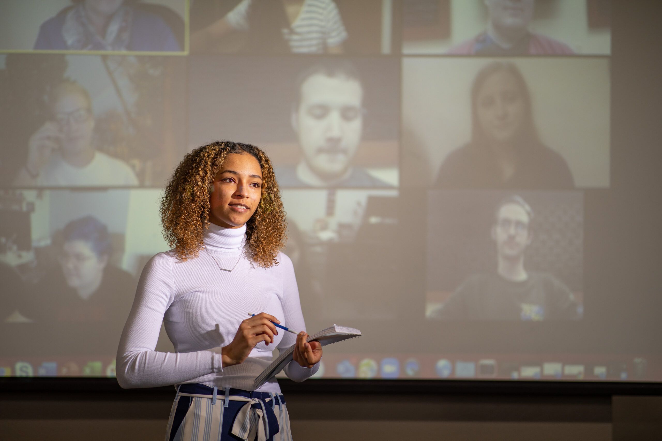 Business student gives a presentation with on-video participants on a projector screen in the background.