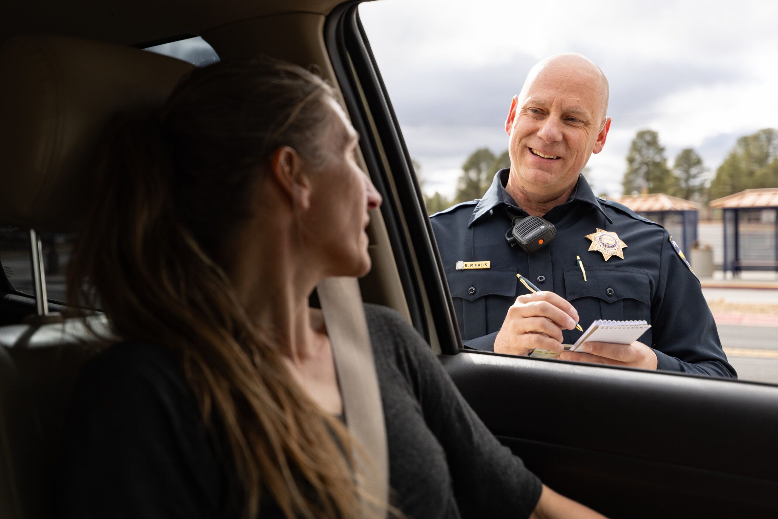 A smiling police officer standing outside a vehicle, speaking with a woman in the driver's seat while taking notes.